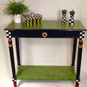 May include: A black and green table with a checkerboard pattern on the legs and a green top. The table has a single drawer with a red and gold knob. There is a green box with a checkerboard pattern and red and green stripes on top of the table. Two black and white checkerboard salt and pepper shakers are on the table.
