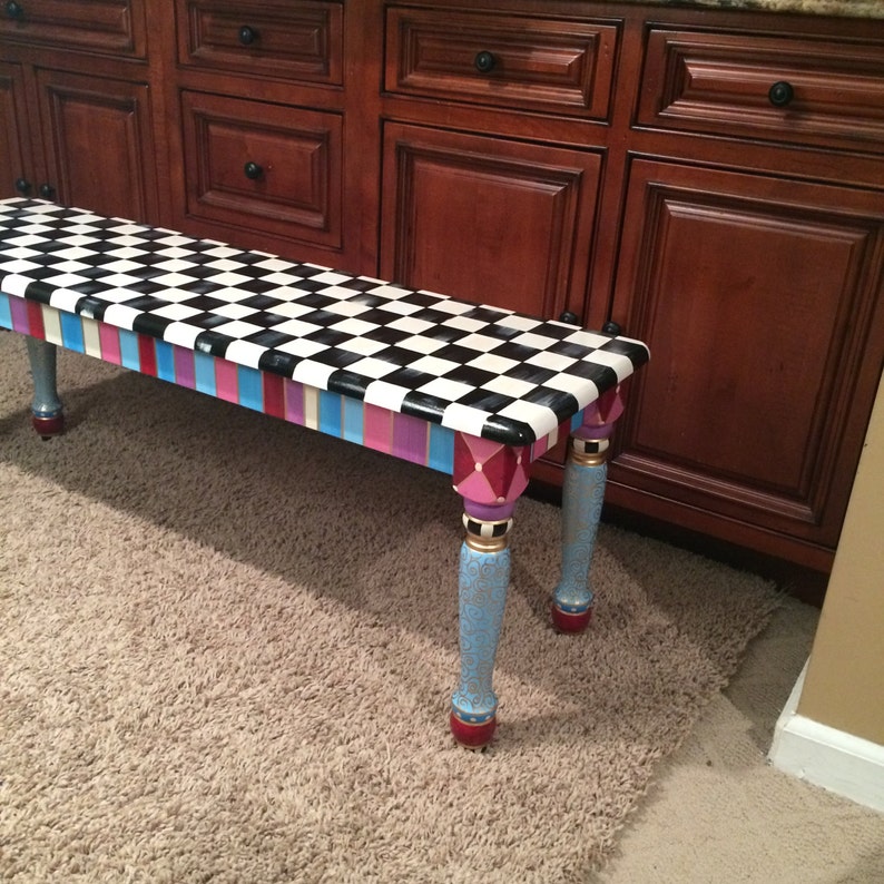 May include: A black and white chequered bench with colourful stripes and diamond patterns on the legs. The bench is painted in a whimsical style and has a blue, pink, and gold colour scheme.