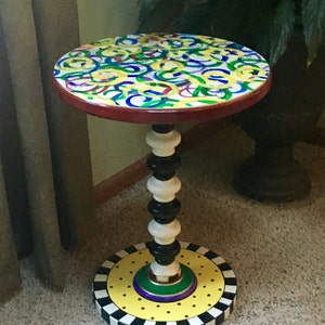 May include: A round side table featuring a tabletop with a colorful pattern of yellow, blue, green, and red swirls. The central pedestal is composed of alternating black and white turned wooden segments, resting on a circular base with yellow dots.