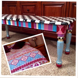 May include: A black and white chequered bench with a colourful striped base. The bench legs are painted with blue, gold, and red accents. The bench is in a room with a patterned bedspread.