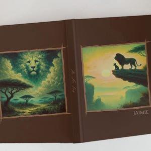 May include: Open journal with a brown cover. The illustrations depict a lion's face in clouds and a lion and cub on a cliff at sunset. The name "Jaimie" is printed on the cover. The text "M. The Lion King" is also visible.