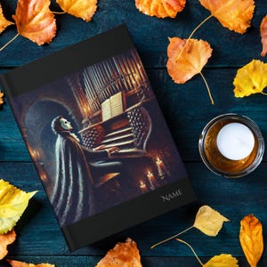May include: A black hardcover journal with a dark, moody image of a masked figure playing a pipe organ in a dimly lit room. The journal has the word "NAME" printed on the front cover.