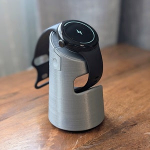 May include: A gray 3D printed charging stand for a black smartwatch with a round face. The watch face shows a lightning bolt icon, indicating it is charging.