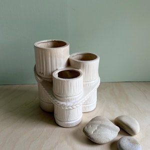 May include: White ceramic three-sectioned planter with a rope design. The planter is shaped like three stacked cylinders. Three smooth, light gray stones are in front of the planter.
