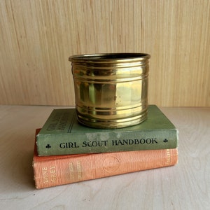 May include: A gold metal planter with a ribbed design sits on top of two vintage books. The top book is green and says "Girl Scout Handbook". The bottom book is orange and says "Zane Grey Thunder Mountain".