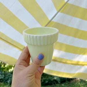 May include: A pale yellow ceramic flower pot with a ribbed rim. The small pot is held against a background of yellow and white striped fabric. The pot is empty and ready for planting.