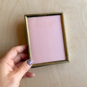 May include: A rectangular, vintage-style photo frame with a gold-colored metal frame and a pink interior. The frame has a decorative, repeating pattern around the edges. The frame is empty and ready for a photo.