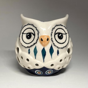 May include: A white ceramic owl-shaped candle holder featuring hand-painted black outlines for the eyes, a light orange beak, and teal diamond-shaped accents on the chest. The base is dark blue with circular patterns and perforated side cutouts.