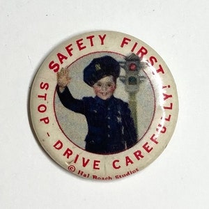 May include: A vintage safety button with a red border and white background. The button features a young boy in a blue uniform holding a traffic light and waving. The text on the button reads "Safety First! Stop - Drive Carefully." and "Hal Roach Studios".