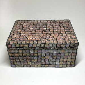 May include: A rectangular box covered in a mosaic of small, square, iridescent tiles. The tiles feature a mix of muted pink, purple, blue, and gold tones with a reflective, pearlescent finish. The box has a flat lid and a structured, boxy shape.