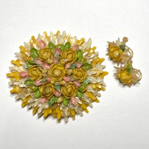 May include: A vintage floral brooch and matching earrings set. The brooch is a circular design with yellow, pink, and green flower details. The earrings have a similar floral design. The set is on a white background.