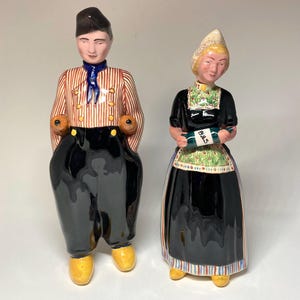 May include: A pair of ceramic figurines depicting a man and a woman in traditional Dutch clothing. The man is wearing a red and white striped shirt, black pants, and a blue cap. The woman is wearing a black dress with a white apron and a yellow and white cap. The woman is holding a bottle labeled "Bols".