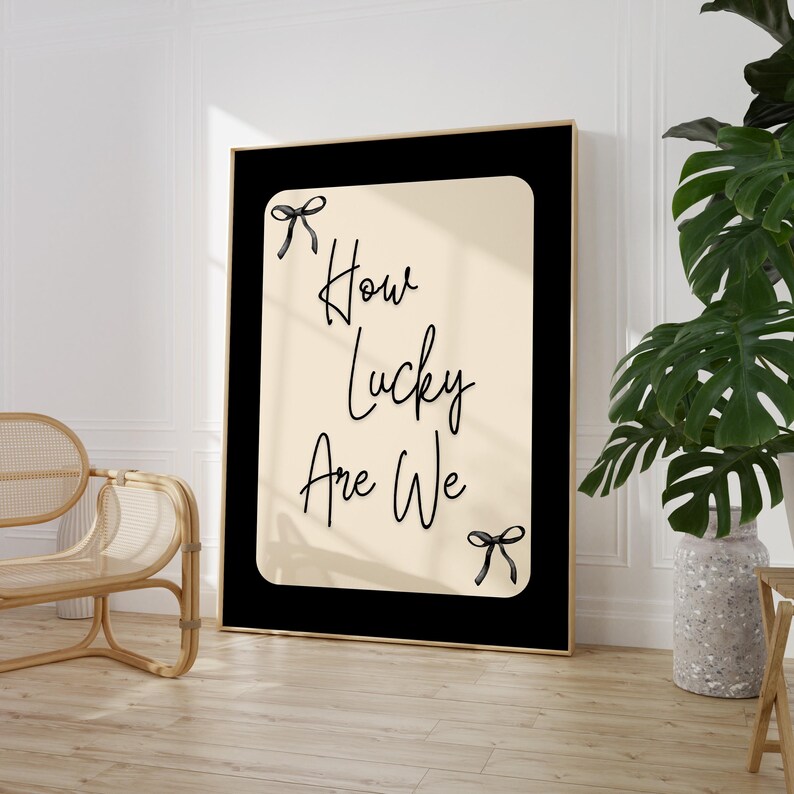 K&ouml;nnte beinhalten: Gerahmter Kunstdruck mit dem Text "How Lucky Are We" in stilvoller Schrift. Das Kunstwerk zeigt einen cremefarbenen Hintergrund mit schwarzem Rand und goldenem Rahmen. Schwarze Schleifenakzente befinden sich oben und unten am Text.