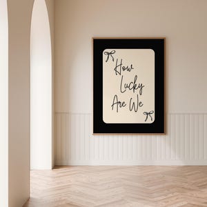 K&ouml;nnte beinhalten: Gerahmter Kunstdruck mit dem Text "How Lucky Are We" in handschriftlichem Stil. Der Druck hat einen schwarzen Rand und einen hellbeigen Hintergrund. Zwei kleine Schleifenillustrationen befinden sich oben und unten. Der Rahmen ist braun.