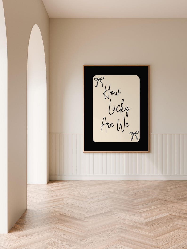 K&ouml;nnte beinhalten: Gerahmter Kunstdruck mit dem Text "How Lucky Are We" in handschriftlichem Stil. Der Druck hat einen schwarzen Rand und einen hellbeigen Hintergrund. Zwei kleine Schleifenillustrationen befinden sich oben und unten. Der Rahmen ist braun.