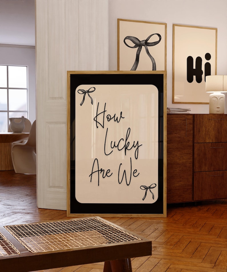 K&ouml;nnte beinhalten: Ein gerahmter Kunstdruck mit dem Text "How Lucky Are We" in schwarzer Schrift, verziert mit kleinen Schleifen-Illustrationen. Das Kunstwerk hat einen cremefarbenen Hintergrund mit einem schwarzen Rand und einem goldenen Rahmen. Weitere gerahmte Kunstwerke sind im Hintergrund zu sehen.