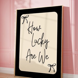 K&ouml;nnte beinhalten: Ein gerahmter Kunstdruck mit dem Text "How Lucky Are We" in eleganter Schrift. Das Kunstwerk zeigt einen cremefarbenen Hintergrund mit schwarzem Text und Schleifen, eingebettet in einen schwarzen Rahmen mit goldenem Rand. Der Druck lehnt an einer rosa Wand.
