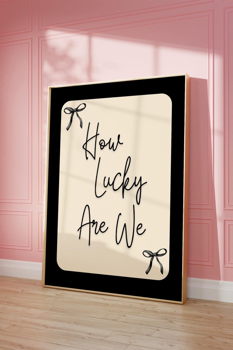K&ouml;nnte beinhalten: Ein gerahmter Kunstdruck mit dem Text "How Lucky Are We" in eleganter Schrift. Das Kunstwerk zeigt einen cremefarbenen Hintergrund mit schwarzem Text und Schleifen, eingebettet in einen schwarzen Rahmen mit goldenem Rand. Der Druck lehnt an einer rosa Wand.
