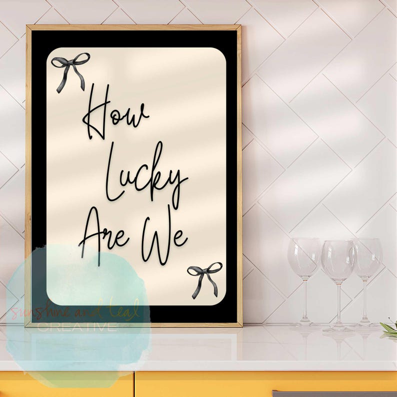 K&ouml;nnte beinhalten: Gerahmter Kunstdruck mit dem Text "How Lucky Are We" in eleganter Schrift, verziert mit schwarzen Schleifen-Illustrationen. Das Kunstwerk hat einen cremefarbenen Hintergrund, gerahmt in Schwarz und Gold, f&uuml;r die Inneneinrichtung.
