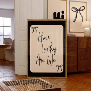 K&ouml;nnte beinhalten: Ein gerahmter Druck mit dem Text "How Lucky Are We" in eleganter Schrift, verziert mit Schleifenillustrationen. Das Kunstwerk hat einen cremefarbenen Hintergrund mit schwarzem Rand und einem goldenen Rahmen. Zus&auml;tzliche gerahmte Kunstwerke sind im Hintergrund zu sehen.