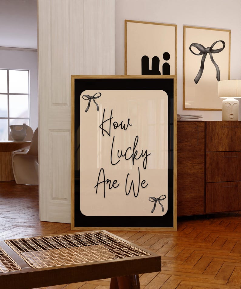 K&ouml;nnte beinhalten: Ein gerahmter Druck mit dem Text "How Lucky Are We" in eleganter Schrift, verziert mit Schleifenillustrationen. Das Kunstwerk hat einen cremefarbenen Hintergrund mit schwarzem Rand und einem goldenen Rahmen. Zus&auml;tzliche gerahmte Kunstwerke sind im Hintergrund zu sehen.