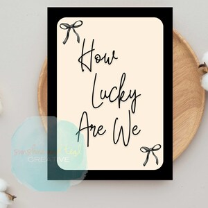 K&ouml;nnte beinhalten: Rechteckige Karte mit schwarzem Rand und dem Text "How Lucky Are We" in schwarzer Schrift. Die Karte hat einen cremefarbenen Hintergrund und ist mit schwarzen Schleifenakzenten verziert.