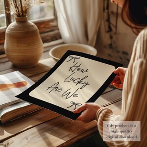 K&ouml;nnte beinhalten: Ein schwarz gerahmter Druck mit dem Text "How Lucky Are We" in eleganter Schrift, verziert mit Schleifenakzenten. Der Druck hat einen cremefarbenen Hintergrund und wird vor einem Holztisch gehalten. Das Bild enth&auml;lt auch eine Vase, eine Sch&uuml;ssel und ein offenes Buch.