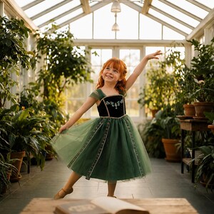 May include: A young girl with red hair wearing a green and black dress with floral embroidery, posing in a greenhouse. The dress has a tulle skirt and short sleeves. The girl is smiling and has one arm raised. The background features lush greenery and sunlight.