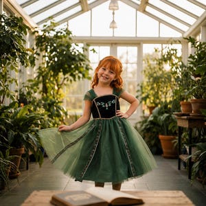 May include: A young girl with red hair smiles, wearing a green and black dress with floral embroidery. The dress has a tulle skirt and short sleeves. She is standing in a greenhouse filled with plants, with a wooden table and an open book in the foreground.