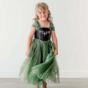 Peut inclure: Une jeune fille porte une robe verte et noire avec un motif floral sur le corsage. La robe a une jupe à plusieurs épaisseurs et des manches courtes. La fille sourit devant un fond blanc. Costume pour enfant.