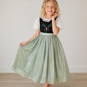 May include: A young girl wearing a dress with a black bodice, white puff sleeves, and a light green skirt. The bodice has floral embroidery. The skirt is a shiny, pleated fabric. The girl is smiling and holding the skirt.