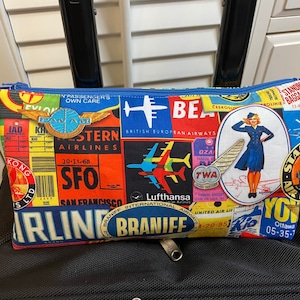 May include: A blue and red zippered pouch with a vintage airline theme. The pouch features logos and text from airlines such as Pan Am, TWA, Braniff, and Lufthansa. The pouch also features a vintage illustration of a flight attendant.