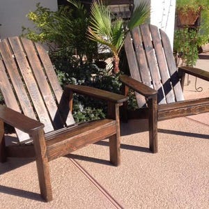 May include: Two wooden Adirondack chairs with a dark brown stain. The chairs have wide armrests and a slatted back design. They are placed on a concrete surface, with greenery in the background.