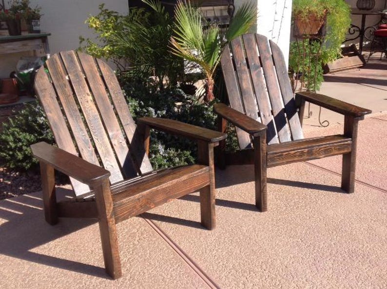 May include: Two wooden Adirondack chairs with a dark brown stain. The chairs have wide armrests and a slatted back design. They are placed on a concrete surface, with greenery in the background.