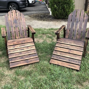 May include: Two wooden Adirondack chairs with a dark brown stain are positioned on a grassy lawn. The chairs have a slatted seat and back, with armrests. The wood grain is visible, and the chairs appear to be handmade.