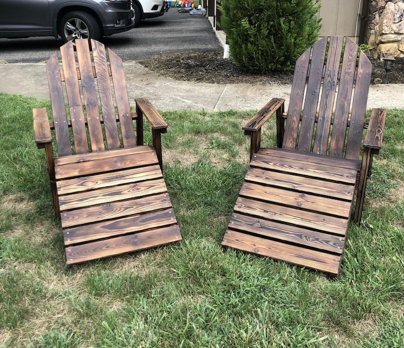 May include: Two wooden Adirondack chairs with a dark brown stain are positioned on a grassy lawn. The chairs have a slatted seat and back, with armrests. The wood grain is visible, and the chairs appear to be handmade.