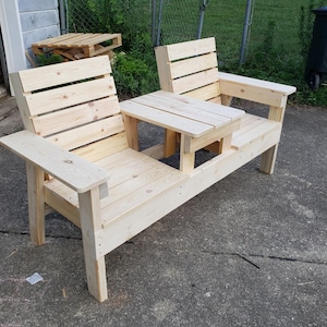 May include: A wooden bench with a built-in table in the center. The bench is made of unfinished pine wood and has two seats. The table is also made of unfinished pine wood and has a rectangular shape.