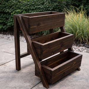 May include: A three-tiered, dark brown wooden planter. The rectangular boxes are stacked, supported by angled side supports and vertical legs. The stained wood has visible grain, suggesting a natural finish. The planter is designed for outdoor use.