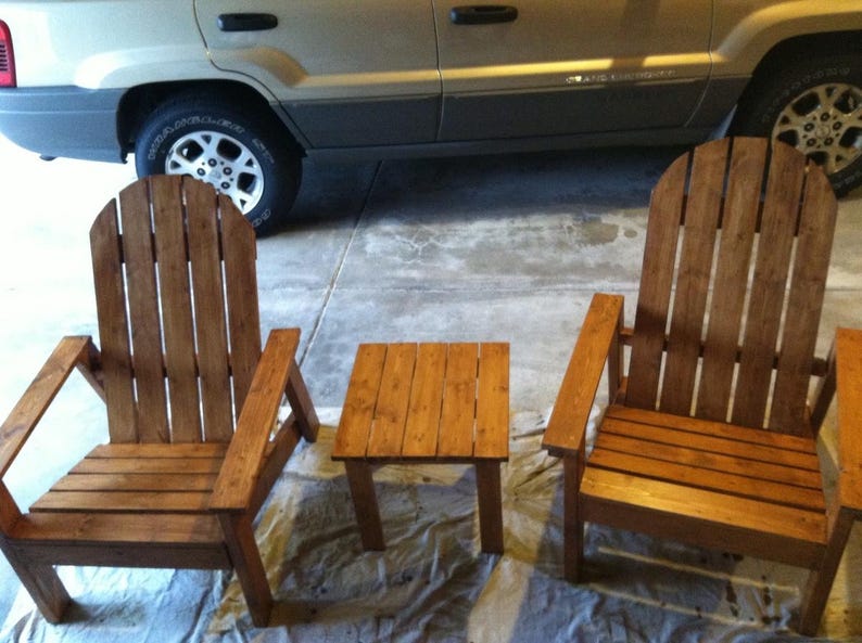 May include: Two wooden Adirondack chairs and a small square side table. The chairs have a slatted back and armrests. The furniture is made of stained wood and is suitable for outdoor use.