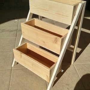 Peut inclure: Un support de jardini&egrave;re en bois &agrave; trois niveaux avec une finition en bois naturel. Le support comporte trois bo&icirc;tes rectangulaires soutenues par un cadre blanc, id&eacute;al pour exposer des plantes ou des fleurs. Le support mesure environ 76 cm de haut.