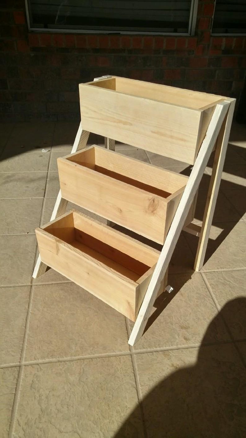 Peut inclure: Un support de jardini&egrave;re en bois &agrave; trois niveaux avec une finition en bois naturel. Le support comporte trois bo&icirc;tes rectangulaires soutenues par un cadre blanc, id&eacute;al pour exposer des plantes ou des fleurs. Le support mesure environ 76 cm de haut.
