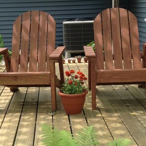 May include: Two wooden Adirondack chairs with a reddish-brown finish sit on a wooden deck. A terracotta pot with red flowers is placed between the chairs. The background includes a blue house and an air conditioning unit.
