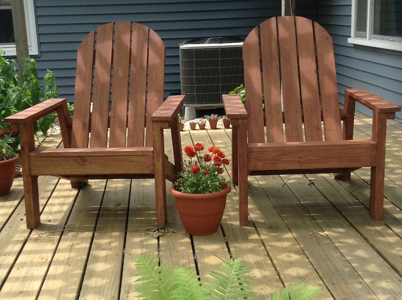 May include: Two wooden Adirondack chairs with a reddish-brown finish sit on a wooden deck. A terracotta pot with red flowers is placed between the chairs. The background includes a blue house and an air conditioning unit.