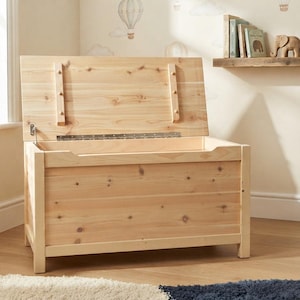 Puede incluir: Un baúl de juguetes rectangular de madera clara con una tapa abatible que se mantiene abierta. El baúl presenta un acabado de madera natural con nudos visibles, una bisagra de piano de metal y un detalle de asa recortada en el panel frontal.