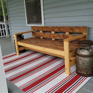 May include: A wooden bench with a rustic finish sits on a porch with a red and white striped rug. A vintage milk can sits next to the bench.