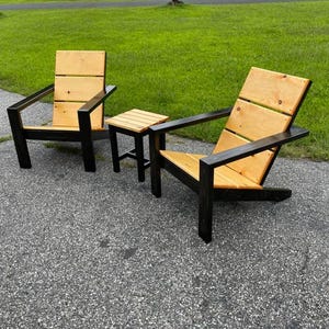 Könnte beinhalten: Zwei Adirondack-Stühle aus Holz und Schwarz mit einem passenden Beistelltisch aus Holz und Schwarz. Die Stühle und der Tisch stehen auf einer grauen Kiesauffahrt.