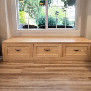 Puede incluir: Un banco de madera de color claro con tres cajones, cada uno con un tirador negro. El banco est&aacute; situado debajo de una ventana, con una vista de vegetaci&oacute;n visible a trav&eacute;s del cristal. El banco es de madera.