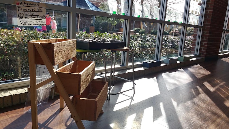 Peut inclure: Un bac &agrave; plantes en bois &agrave; trois niveaux avec trois bo&icirc;tes rectangulaires. La jardini&egrave;re est plac&eacute;e pr&egrave;s d'une grande fen&ecirc;tre, avec la lumi&egrave;re du soleil qui entre. Un panneau est visible en arri&egrave;re-plan. D'autres jardini&egrave;res sont sur un tabouret et un rebord de fen&ecirc;tre.