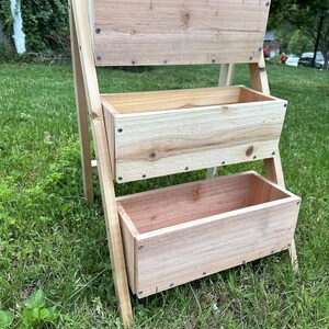 Peut inclure: Un jardini&egrave;re en bois &agrave; trois niveaux avec une finition naturelle. La jardini&egrave;re comporte trois bo&icirc;tes rectangulaires pour la plantation, soutenues par une structure en forme de A. Le grain du bois est visible, et la jardini&egrave;re est &agrave; l'ext&eacute;rieur sur une surface herbeuse.