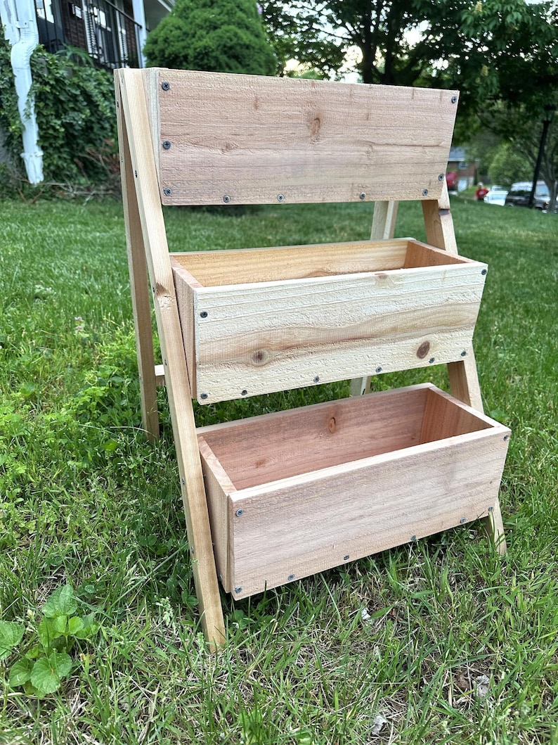 Peut inclure: Un jardini&egrave;re en bois &agrave; trois niveaux avec une finition naturelle. La jardini&egrave;re comporte trois bo&icirc;tes rectangulaires pour la plantation, soutenues par une structure en forme de A. Le grain du bois est visible, et la jardini&egrave;re est &agrave; l'ext&eacute;rieur sur une surface herbeuse.